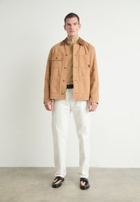 Tanskleurige katoen jas met vier zakken, bruin kraag en knopen, gedragen over een beige zip-up top, gecombineerd met witte broeken en zwarte schoenen.