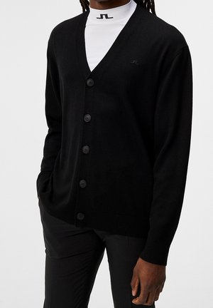 Man die een zwarte, dichtgeknoopte cardigan draagt over een wit shirt met hoge hals en een zwart logo, gecombineerd met een zwarte broek, staand met één hand in de zak.