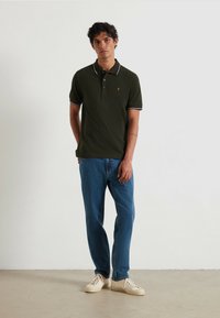 Polo shirt verde scuro con bordi bianchi, patta con tre bottoni e piccolo logo ricamato, abbinato a jeans in denim blu e sneakers crema.
