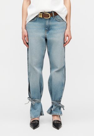 Ανοιχτό μπλε τζιν με πλαϊνές σχισμές και δέσιμο στους αστραγάλους, φορεμένο με λευκό τοπ, καφέ ζώνη και μαύρα παπούτσια με μυτερή μύτη.