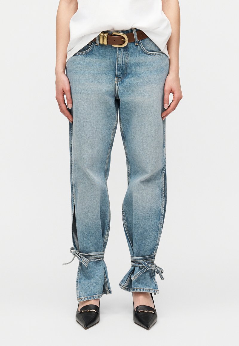 Ανοιχτό μπλε τζιν με πλαϊνές σχισμές και δέσιμο στους αστραγάλους, φορεμένο με λευκό τοπ, καφέ ζώνη και μαύρα παπούτσια με μυτερή μύτη.