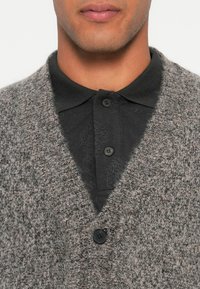 Mand iført en mørkegrå polo shirt lagdelt under en tyk, grå strikket cardigan med en stor sort knap.