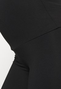 Detailní záběr na černé legíny s vysokým pasem, které ukazují hladkou, elastickou tkaninu a detaily švů podél boků a pasu.