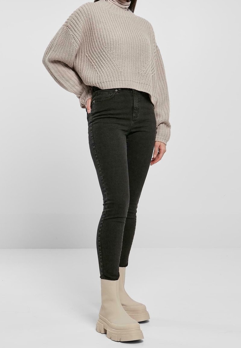 Beige gebreide trui, zwarte high-waisted skinny jeans en crèmekleurige enkellaarsjes met stevige zolen. Soepele textuur en relaxed fit.