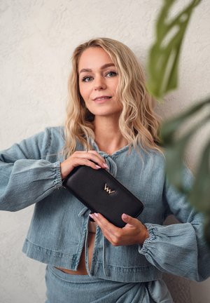 Black leather wallet with gold accent, rectangular shape, featuring a zipper closure and embossed logo, held by a person in a denim outfit.