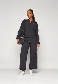 Μαύρο κοστούμι γυμναστικής με λευκές πλευρικές ρίγες, που διαθέτει μπουφάν με φερμουάρ και χαλαρούς παντελόνια. Συνδυάζεται με λευκά αθλητικά παπούτσια και μια μικρή μαύρη τσάντα.