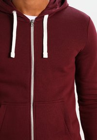Sudadera con cremallera en burdeos, hecha de tela suave, con cordón blanco, bolsillos frontales y una cremallera plateada suave.