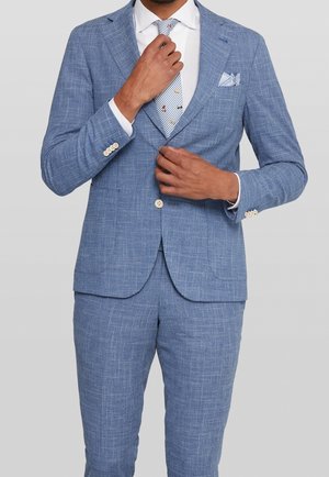Hombre ajustando chaqueta de traje azul claro texturizada y pantalones a juego con camisa blanca y corbata estampada sobre fondo gris.