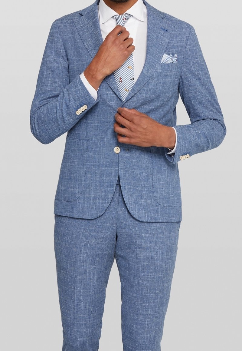 Homme ajustant une veste de costume bleu clair texturée et un pantalon assorti avec une chemise blanche et une cravate à motifs sur fond gris.
