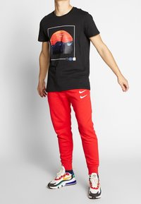 Svart grafisk t-shirt med en röd och blå bergsprint, tillsammans med röda Nike sweatpants och multikoloriga träningsskor.