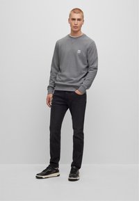 Uomo in felpa grigia, jeans neri e sneaker nere in piedi davanti a uno sfondo grigio chiaro, con una mano in tasca.