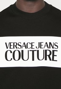 Černá mikina s výrazným bílým horizontálním pruhem a textem "VERSACE JEANS COUTURE" v černém písmu. Žebrovaný výstřih.