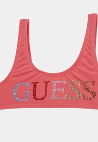 Top sportivo rosa corallo con spalline larghe, caratterizzato da lettere in strass multicolori che scrivono "GUESS" sulla parte anteriore. Texture del tessuto liscia.