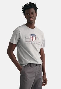 Grijze katoenen t-shirt met een rood en blauw schildlogo met de tekst "GANT" en "AMERICAN SPORTSWEAR", ronde halslijn en korte mouwen.