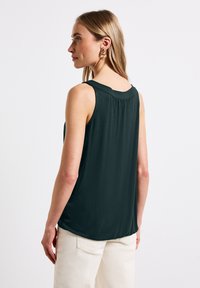 Top senza maniche di un verde scuro con scollatura arrotondata, morbida al tatto, e dettaglio di drappeggio sul retro; indossato con pantaloni beige chiaro.