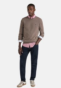 Maglione marrone sopra una camicia rosa chiaro con colletto, abbinato a jeans di denim scuro e sneakers beige, con un logo sul maglione.
