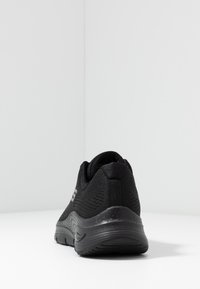 Svart sportssko med ett texturerat meshöverdel och en vadderad häl, med en slank, rund sula och minimalistisk branding i silver.