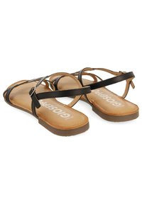 Sandalias de cuero negro con tiras cruzadas, una plantilla beige y una suela plana marrón. Presenta un detalle de hebilla en el lateral para la ajustabilidad.