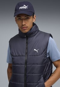 Granatowa kamizelka puchowa z wysokim kołnierzem, zamkiem b łyskawicznym i białym logo na piersi; noszona na jasnoniebieskiej koszulce z krótkim rękawem i granatowej czapce.