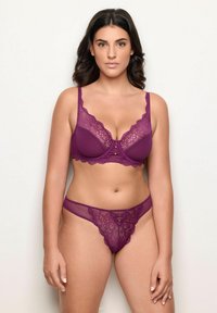 Femme portant un ensemble de lingerie en dentelle violette assorti, se tenant contre un fond clair uni, faisant face à l'avant avec une expression neutre.