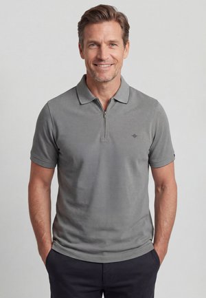 Lächelnder Mann mit kurzen Haaren, der ein graues kurzärmeliges Polo mit Reißverschluss und dunkle Hose trägt, Hände in den Taschen, vor hellem Hintergrund.