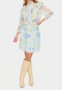 Häikäisevä, pastellivärinen kukkamekko, jossa on frilloja ja nappisuljenta, smockattu vyötärö ja liehuva helma. Yhdistetty polvipitkäisiin beigeihin saappaisiin.