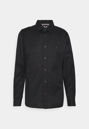 Chemise noire à manches longues, boutonnée, avec col et petit logo sur la poitrine gauche, sur fond blanc uni.