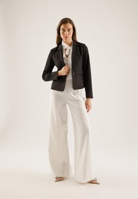 Schwarzer maßgeschneiderter Blazer mit Knopf, kombiniert mit weißen weit geschnittenen Hosen. Weiße Bluse mit Bindeband. Glatter Stoff, strukturiertes Design.