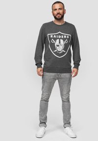 Grauer Sweatshirt mit weißem "Raiders"-Grafik und Logo. Gerippte Bündchen und Saum. Getragen mit hellgrauen Slim-Fit-Jeans und weißen Sneakers.