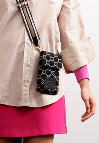 Schwarze Stoff-Handytasche mit weißen Mustern, ausgestattet mit einem Klappenverschluss und einem Logopatch, der an einem gestreiften Trageriemen befestigt ist. Wird über einer hellen Bluse getragen.