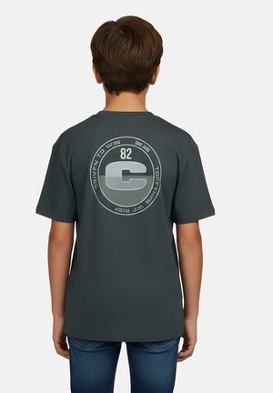 Jongen gezien van achteren, draagt een donkergrijs T-shirt met een cirkelvormig logo met het nummer "82" en de tekst "Driven to win" en "Together we rise".