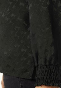 Schwarzes Oberteil mit einem strukturierten Muster, das am Armabschluss mit einem gerafften Smockingdetail versehen ist. Hergestellt aus einem leichten, leicht durchsichtigen Stoff.