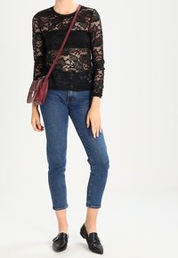 Top lungo a maniche in pizzo nero con motivo floreale, abbinato a jeans blu a vita alta e mocassini neri. La borsa a tracolla bordeaux aggiunge un contrasto.