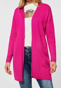 Cardigan long fuchsia à devant ouvert, manches côtelées, et texture douce, superposé à une chemise blanche avec un motif pailleté doré.