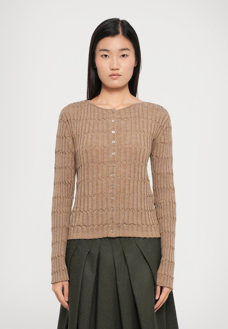 Light brown knitted cardigan with a ribbed texture, round neckline, and button closure, paired with a dark green pleated skirt.
