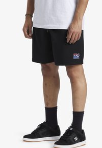 Pantaloni sportivi neri con un motivo testurizzato, con logo DC Athletics sull'orlo inferiore. Abbinati a scarpe da ginnastica nere e calze corte.