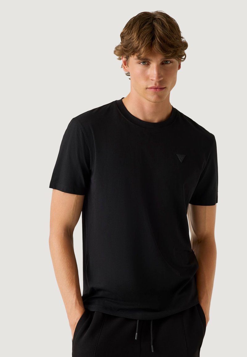 Jeune homme aux cheveux châtain clair portant un t-shirt noir uni à manches courtes avec un col rond et un pantalon noir, les mains dans les poches.