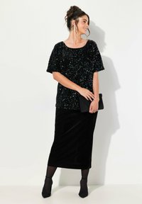 Černý top s flitry a krátkými rukávy, kombinovaný s černou sametovou sukní. Modelka drží černou kabelku; černé punčocháče a lodičky dokončují outfit.