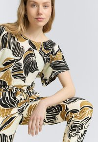 Gemustertes Zweiteiler-Set bestehend aus einem kurzärmeligen Oberteil und passenden Hosen. Das Design umfasst schwarze, beige und senfgelbe Blättermuster auf einem hellen Hintergrund.