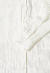 Bílá texturovaná košile na knoflíky s žebrovanou látkou, s manžetou a pěti knoflíky na přední části, která ukazuje jemný, pomačkaný design.