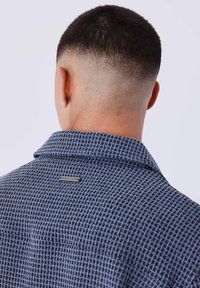 Camicia blu navy con trama a quadretti, dotata di colletto e di un'etichetta in metallo sulla parte superiore della schiena. Capelli corti e tagliati visibili.