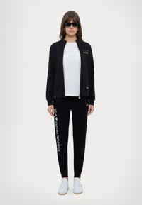 Svart zip-up jacka med gulddetaljer, vit t-shirt, svarta joggers med logotyp och vita sneakers; modell som bär solglasögon.