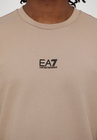 Beige színű pamut póló kerek nyakkivágással. Fekete nyomott "EA7 EMPORIO ARMANI" logó a középen, sima tapintással.