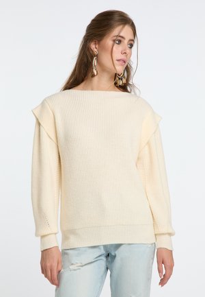 Maglione a maglia color crema con ampio scollo a barca, texture a costine e maniche a campana, indossato sopra jeans chiari strappati.