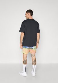 Sort løstsiddende t-shirt, mønstrede mesh-shorts i pastelfarver, hvide sneakers og knælange sokker med sorte striber. Tatoveringer synlige.