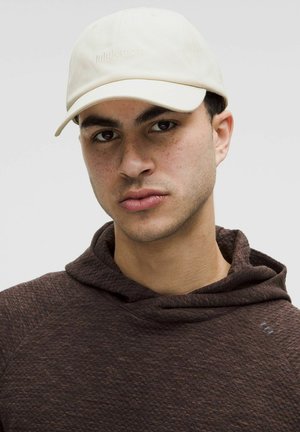 Casquette de baseball blanche avec un logo embossé, portée au-dessus d'un sweat à capuche texturé marron foncé. Le modèle a les cheveux courts et des taches de rousseur visibles.