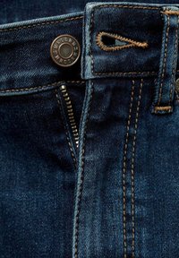 Jean en denim bleu foncé avec une fermeture éclair ouverte, un bouton en métal gravé avec du texte et des coutures marron visibles autour de la ceinture et de la boutonnière.