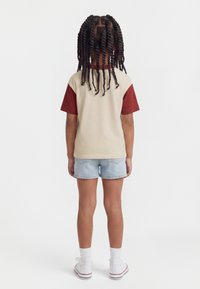 Παιδί με μακριά πλεξούδα, φορώντας μπλούζα κοντομάνικη σε μπεζ και μαρόν χρώμα, denim σορτς, λευκές κάλτσες και λευκά αθλητικά παπούτσια, στέκεται με πλάτη προς τα εμπρός.