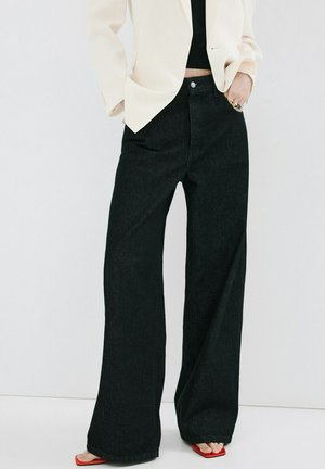 Mujer con vaqueros negros de pierna ancha, un blazer color crema, top negro, tacones rojos abiertos y anillos en la mano derecha.