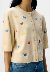 Pull beige à manches courtes, mettant en avant des broderies de papillons multicolores en jaune, bleu, rouge et marron. Design à boutons sur le devant.